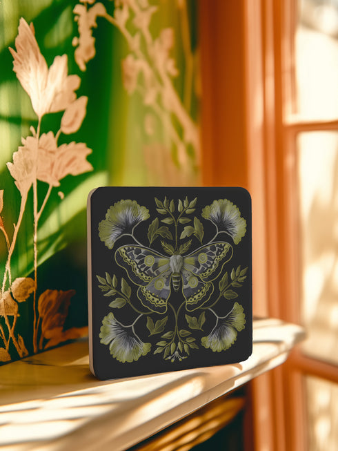 Square black ceramic coaster featuring a detailed chartreuse luna moth illustration surrounded by symmetrical botanical elements including ginkgo-inspired leaves and delicate foliage.